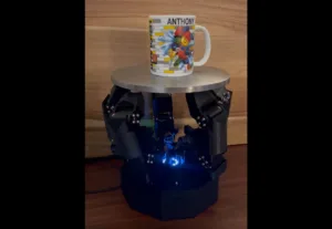 Ceramic mug with colorful LEGO-style artwork sits on a round metal platform atop a black robotic base with blue LEDs visible underneath.