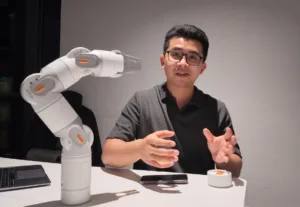 A man wearing glasses sits at a white table while a large white robotic arm extends toward him, explaining or demonstrating something.