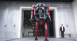 Large red robotic exoskeleton on a wheeled base emerging from a building entrance, with a man in a suit standing nearby.