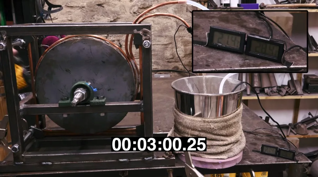 Lab bench with copper coil inside a metal frame and a digital timer displaying 00:03:00.25; cluttered workshop in background.