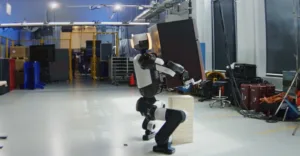 Two‑legged humanoid robot with white and black segments stands on a wooden block in a spacious lab with cables and equipment around.