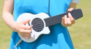 Person in a blue dress plays a small white ukulele outdoors, focusing on the strings and neck.