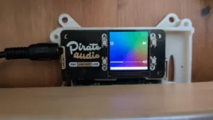 Pirate Audio module with a small color LCD showing music playback, mounted in a white enclosure.