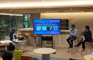 Three people sit on high stools giving a presentation in a modern office, a large screen shows slides behind them while one holds a microphone.