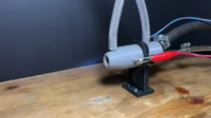 Gray cylindrical hose connector mounted on a black metal bracket with braided hoses and a red clip attached to a wire on a wooden workbench.