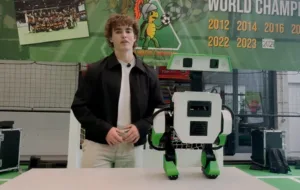 Young man in a black jacket explains a white-and-green two‑legged robot at a robotics exhibit, with a World Championship banner in the background.