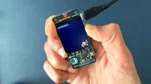 Handheld electronic prototype with a blue screen reading 'LISTENING...' and a cable connected at the top, held in two hands against a blue background.