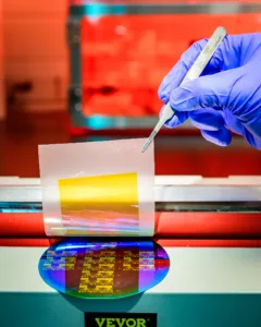 Gloved hand holding a metal lab instrument over a transparent sample pouch in a bright lab setting with red equipment in the background.