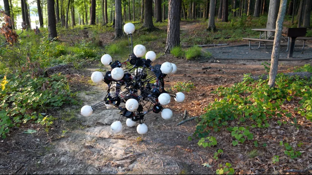 Abstract spherical sculpture of white balls on a dark metal framework in a forest clearing near a picnic table in the background.