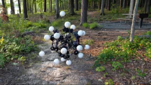 Abstract spherical sculpture of white balls on a dark metal framework in a forest clearing near a picnic table in the background.