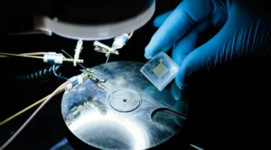 Gloved hand holds a translucent microchip above a metallic circular lab stage with probes nearby.