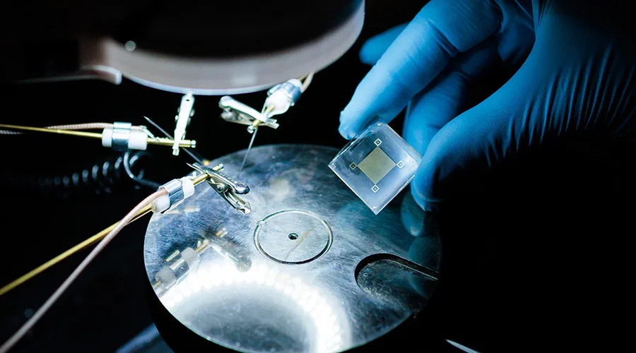 Gloved hand holds a translucent microchip above a metallic circular lab stage with probes nearby.