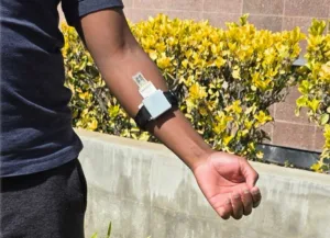 Person's forearm with a white rectangular wearable device strapped on, outdoors near yellow bushes.