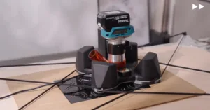 Close-up of a lab bench device: a blue Milwaukee M18 battery atop a metal post, with an orange tube and a black base on a printed mat, wires nearby.