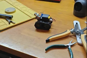 Small DIY tracked robot on a wooden workbench, with pliers and cutters nearby on a yellow work mat and tools in the background.