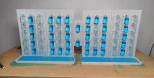Display of blue liquid-filled bottles arranged in vertical columns on white mounting panels with blue trays at the bottom (lab-style setup).