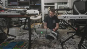 Man with a beard sits on the floor amid keyboards and synthesizers in a home studio, handling a gray drone-like device with green lights.