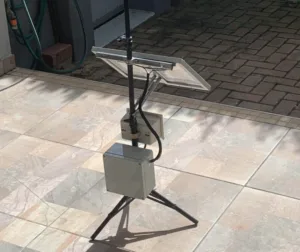 Outdoor tripod-mounted device with a small solar panel and connected electrical boxes on a tiled patio area.