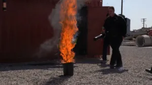 Person in a dark protective uniform stands beside a ground flame, holding a large nozzle or torch near an industrial structure on a gravel lot.