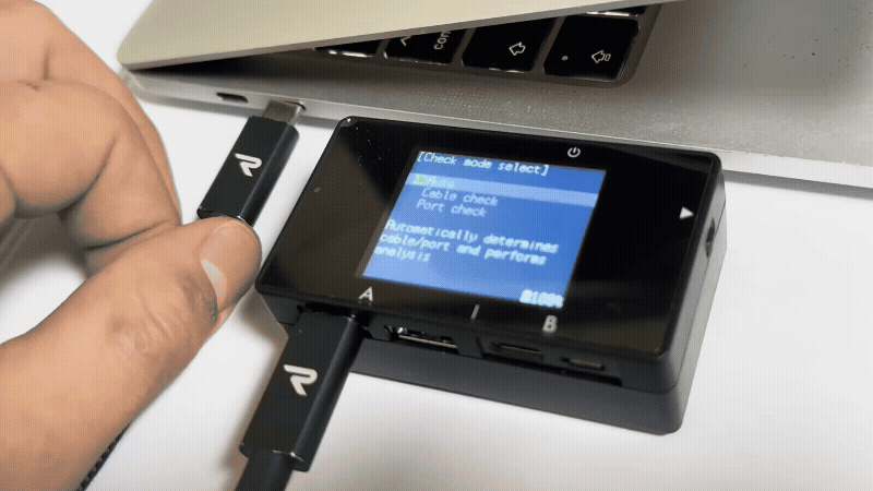 Close-up of a hand inserting a USB-C cable into a USB tester with a blue menu screen under a laptop.