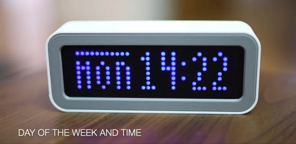 White-framed digital alarm clock with a blue dot-matrix display on a wooden surface, showing the time 10:11.