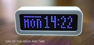 White-framed digital alarm clock with a blue dot-matrix display on a wooden surface, showing the time 10:11.