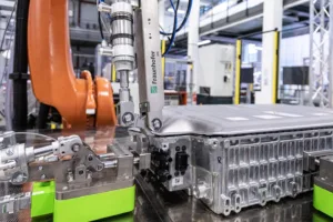 Robot arm with precision gripper assembling a metal enclosure on a factory line, blue cables overhead and orange machinery nearby.
