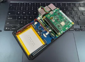 Open handheld electronics prototype on a laptop keyboard, showing a green circuit board, USB ports, and a yellow lithium battery inside a case on the desk.