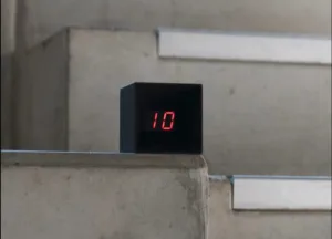 Black cube on a concrete stair displaying red LED number '10' on a stairwell step (floor indicator).