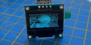 Small OLED display module with four labeled I2C pins (GND, VCC, SCL, SDA) on a blue grid mat.