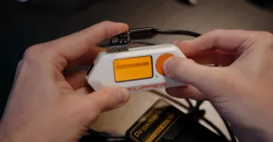 Hands insert a memory card into a small white handheld device with an orange screen and circular orange button labeled 'Clipper'.
