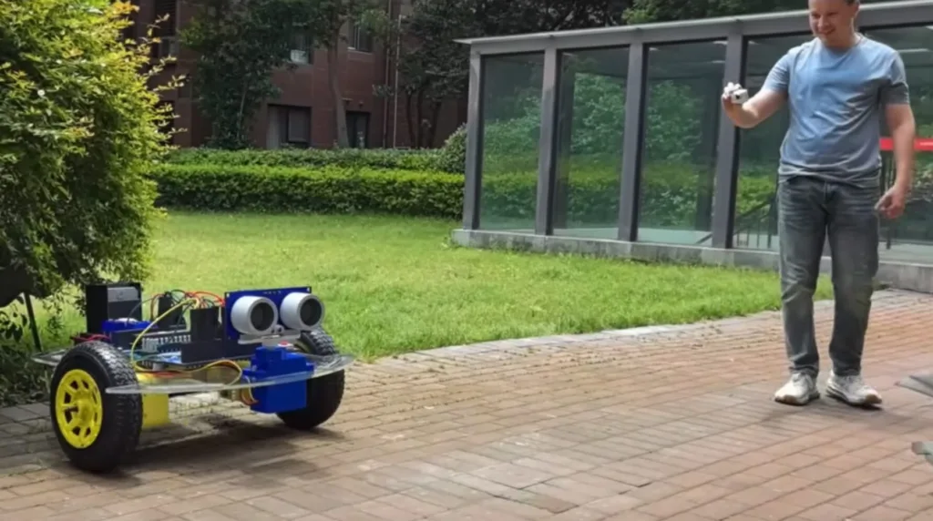Casual outdoor scene with a wheeled robot on a brick path and a man in a blue shirt holding a small object nearby.
