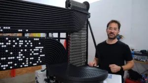 Engineer in a black shirt stands beside a large black modular machine with ribbed panels and rows of white beads on tracks in a lab workshop.