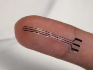 Close-up of a fingertip showing a thin, flexible electronic tattoo with two parallel copper-like lines along the skin and three small black contact pads at the side of the nail.
