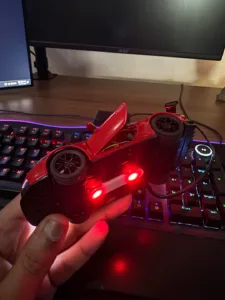 Red toy race car with open cockpit resting on a black game controller, illuminated by red LEDs on a desk with RGB keyboard lighting.
