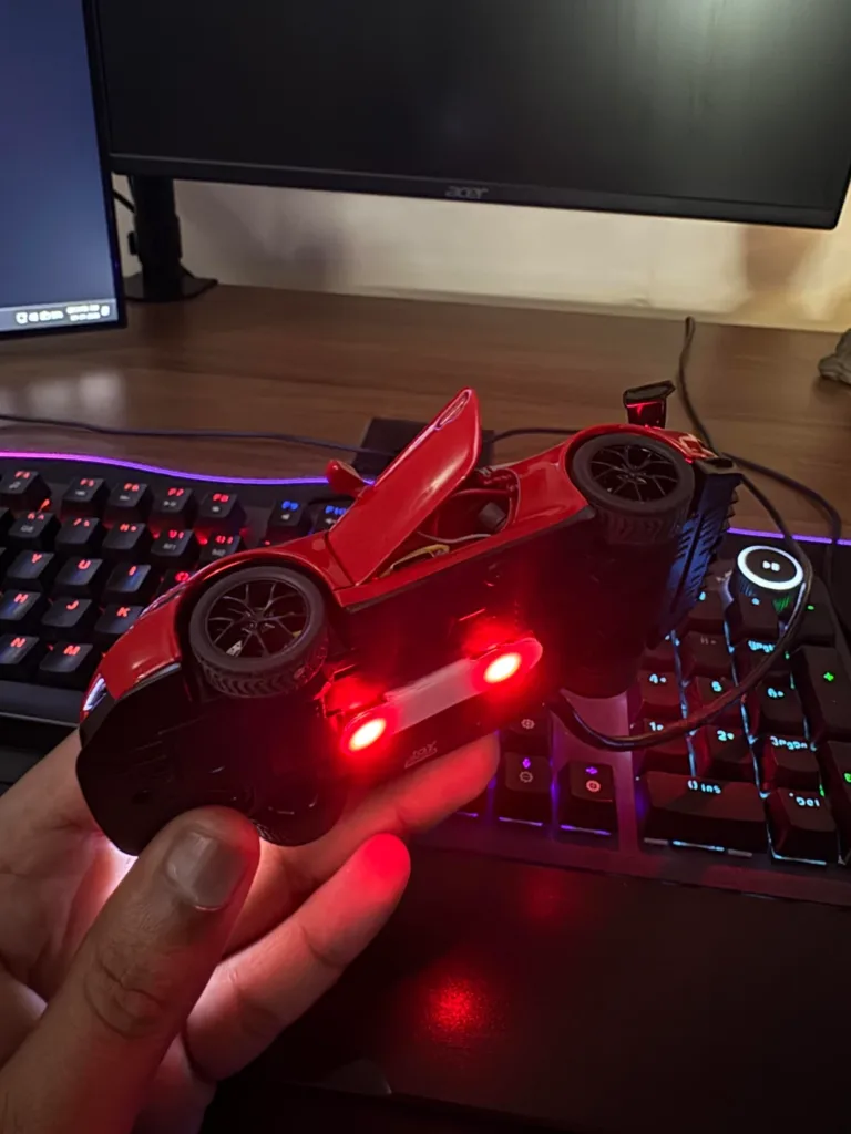 Red toy race car with open cockpit resting on a black game controller, illuminated by red LEDs on a desk with RGB keyboard lighting.