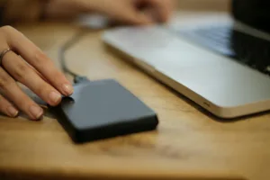 Close-up of a person’s hands using a tablet on a wooden desk with a laptop blurred in the background.