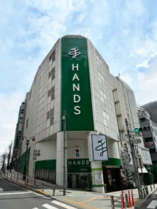 Large green-and-white building with a tall vertical 'HANDS' sign on a city street, Japan, daylight