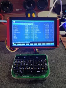 Small handheld computer with a blue terminal window listing files above a compact keyboard on a desk.