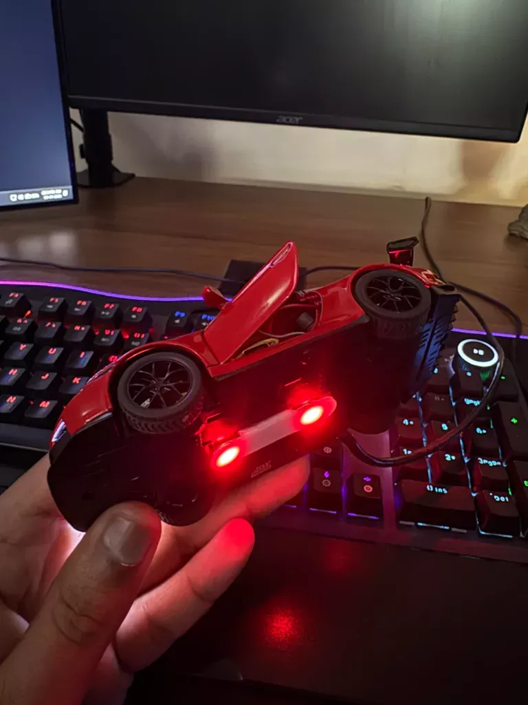 Red toy race car with open cockpit resting on a black game controller, illuminated by red LEDs on a desk with RGB keyboard lighting.