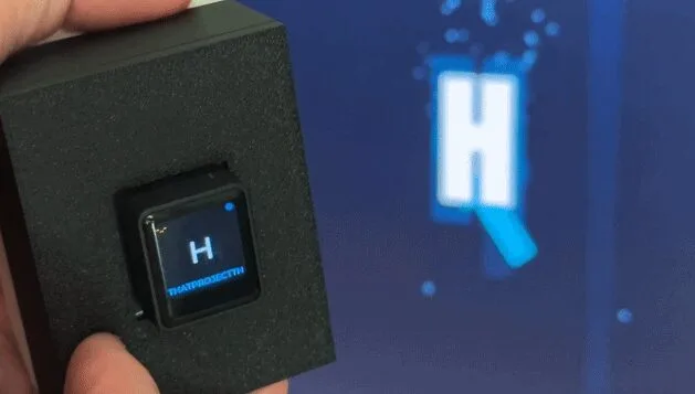 Close-up of a hand holding a small black device with a blue glowing 'H' on its screen; a larger blue 'H' on a screen is blurred in the background.