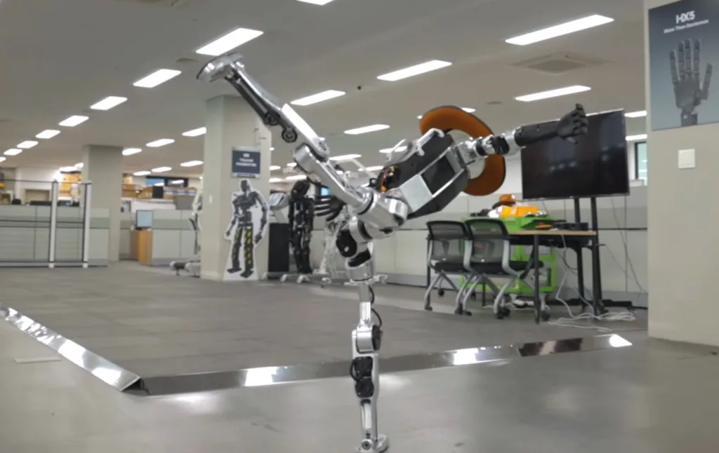Humanoid industrial robot extending its arm in a modern office or lab space with desks and a large monitor behind it.