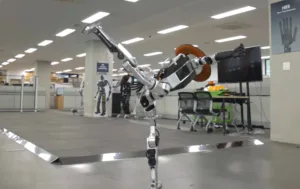 Humanoid industrial robot extending its arm in a modern office or lab space with desks and a large monitor behind it.