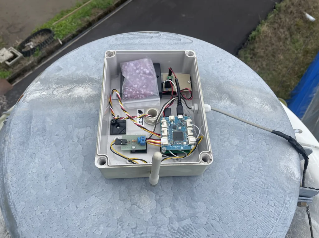 Open plastic enclosure housing a PCB and wiring, mounted on a rounded metal surface outdoors; includes a small bag with pink beads and various connectors.