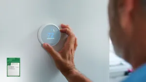Person adjusting a round smart thermostat on a light wall, with a green circuit-board module nearby (installation context)
