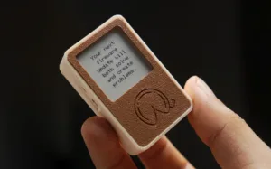 Close-up of a small handheld device with a brown textured front, showing the screen text: "Your next firmware update will both solve and create problems."