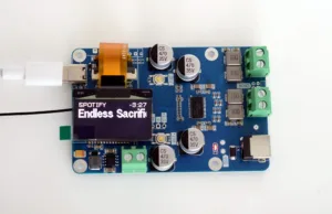 Blue electronic development board with USB port, orange ribbon cable, two large CS 470 35V capacitors, green terminal blocks, and a small display showing 'Spotify Endless Sacrifi' text.