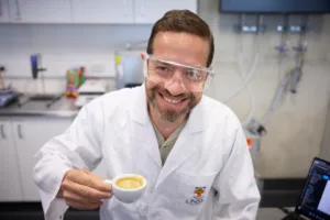 Male scientist in a lab coat and safety goggles smiles while offering a small cup of coffee in a lab setting