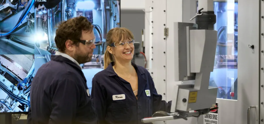 Two engineers in safety glasses and navy coveralls stand beside a large automated machine, smiling in a high-tech lab setting.