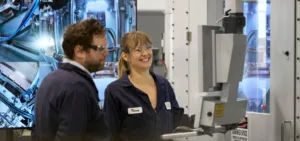 Two engineers in safety glasses and navy coveralls stand beside a large automated machine, smiling in a high-tech lab setting.
